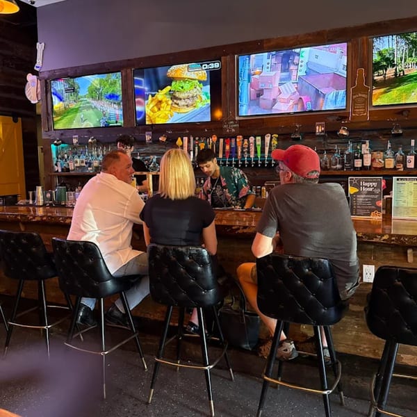 Three patrons sit at a bar with a bartender, watching sports and food on multiple TVs.