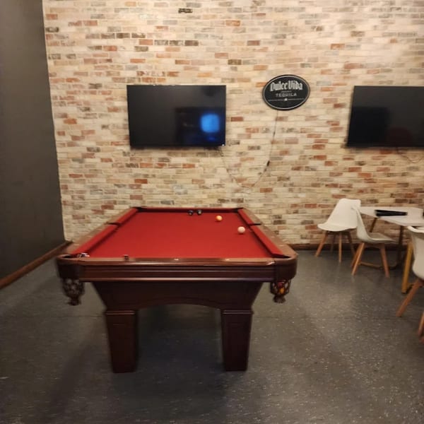 A pool table with red felt and a few balls sits in a room with brick walls and TVs.