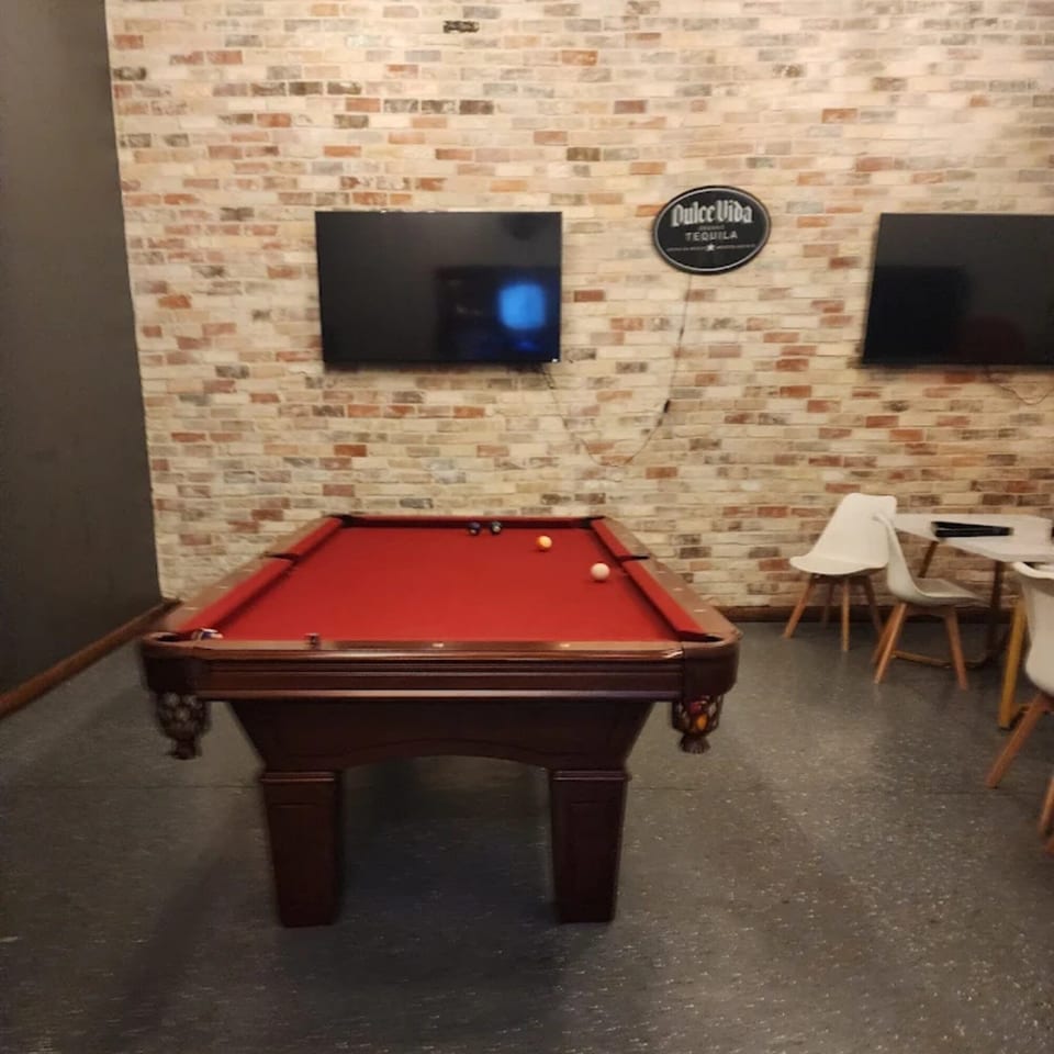 A pool table with red felt and a few balls sits in a room with brick walls and TVs.