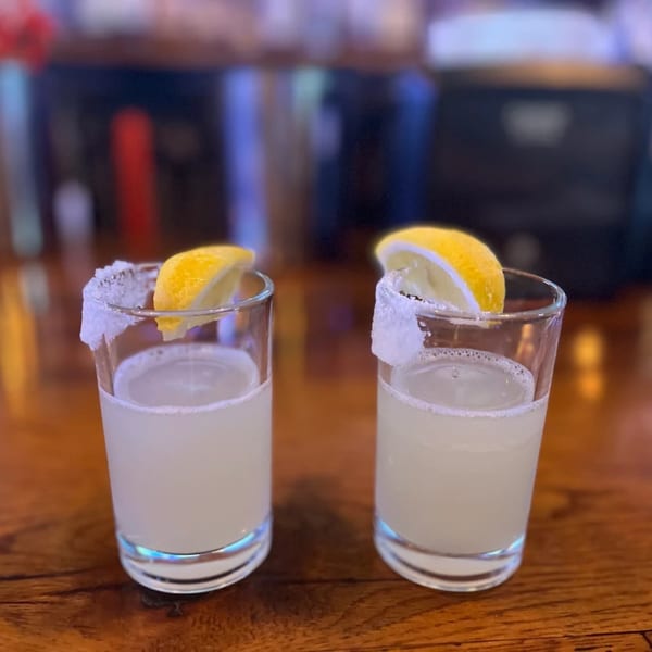 Two lemon drop shots with salted rims and lemon wedges on a wooden bar.
