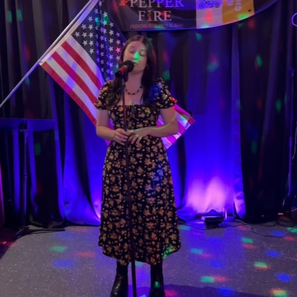 A woman in a floral dress sings into a microphone on a stage with an American flag and colorful lights.