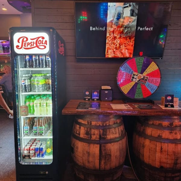 A Pepsi-Cola cooler, a TV showing "Perfect Bulgogi," and a colorful prize wheel on a barrel table.