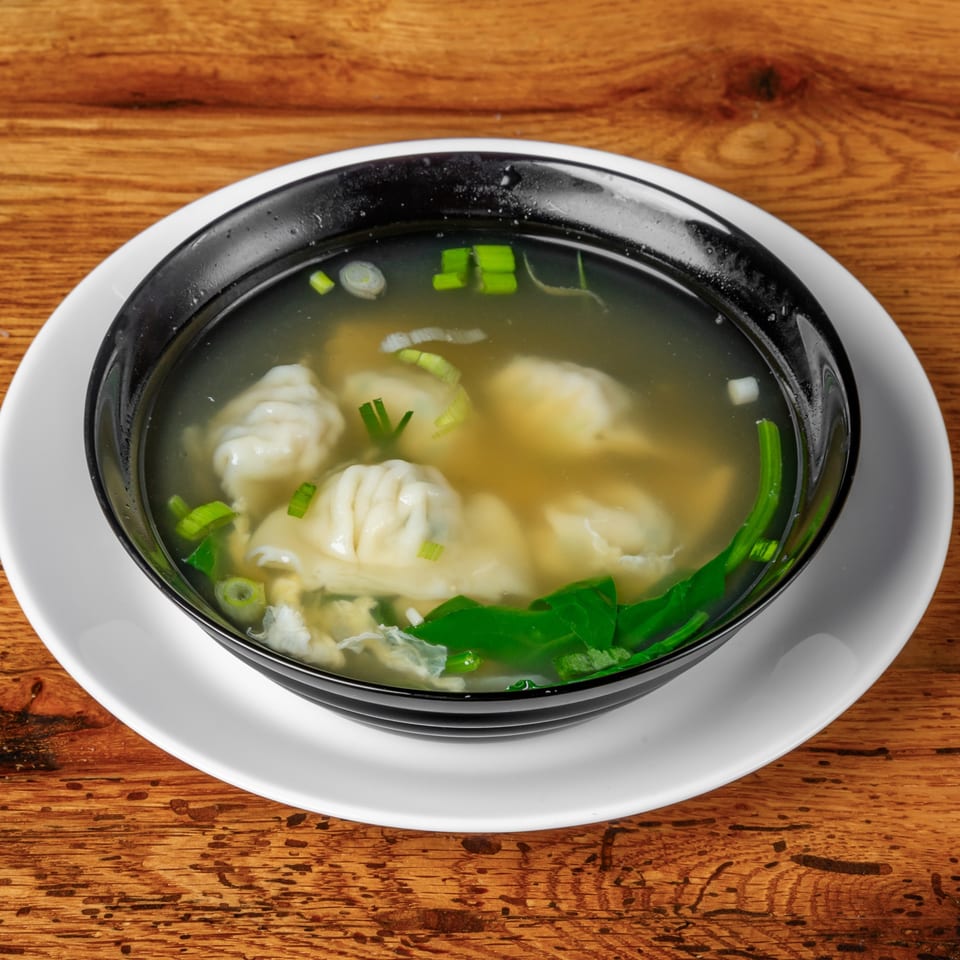 Korean dumpling soup with green onions and egg in a black bowl on a wooden table.