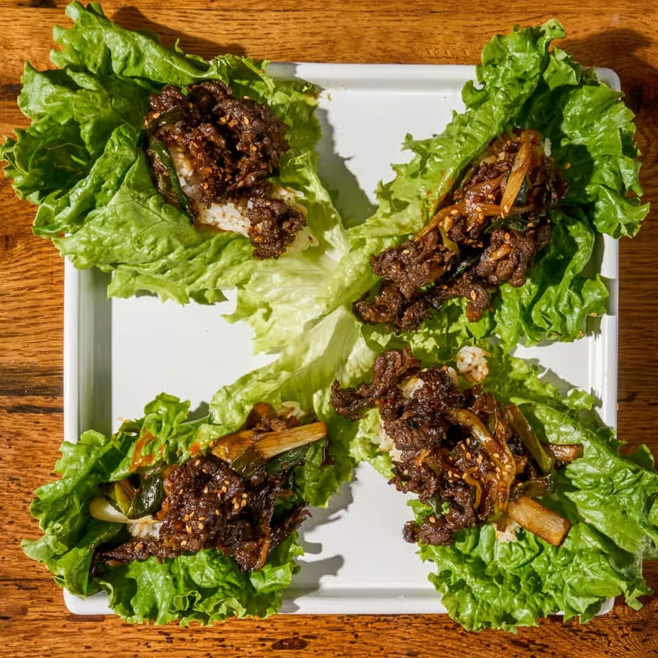 Four Korean beef lettuce wraps with rice, green onions, and sesame seeds on a white plate.