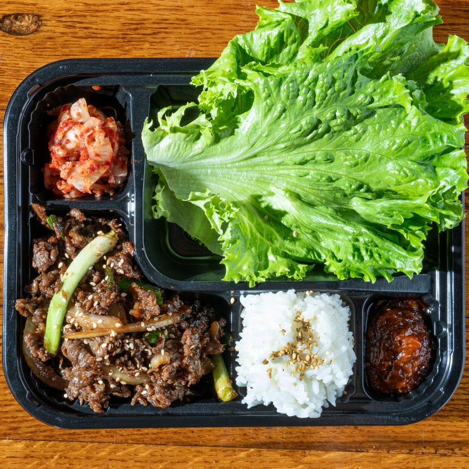 Korean bulgogi meal in a black bento box with rice, kimchi, lettuce, and gochujang.