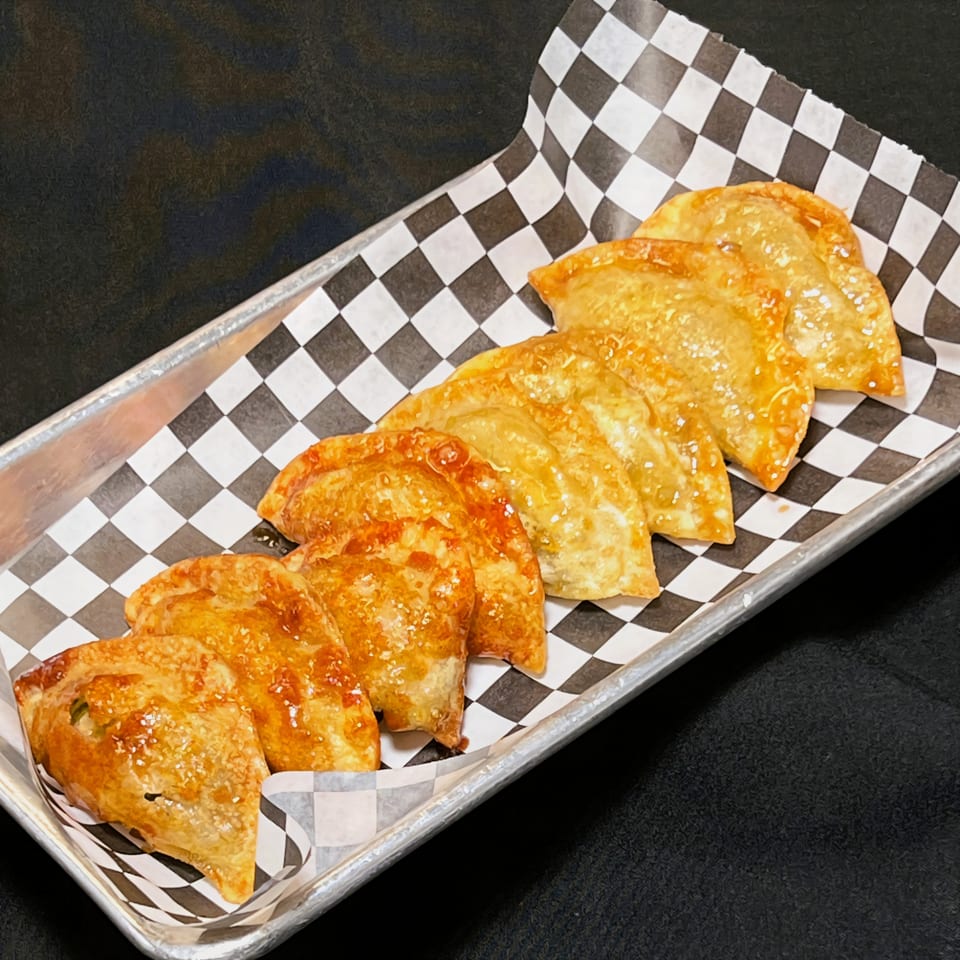 A metal tray holds a line of golden-brown Korean dumplings on black and white checkered paper.