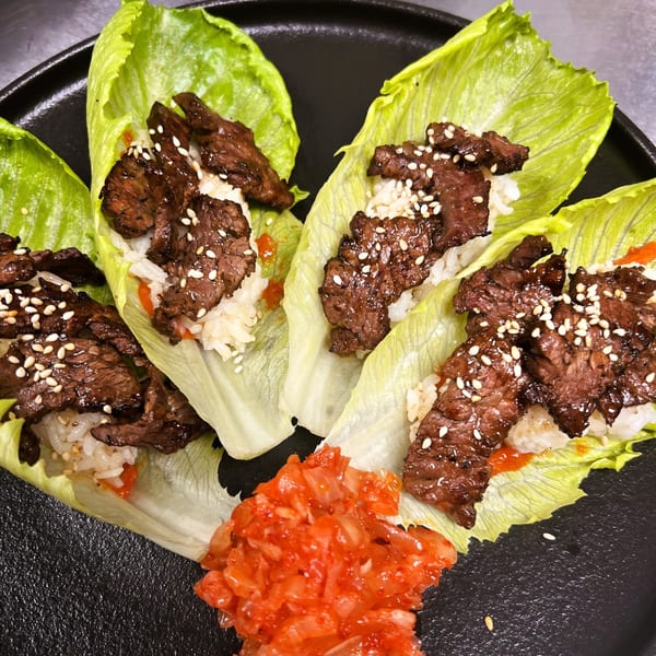 Four lettuce wraps filled with grilled meat, rice, sesame seeds, and a side of kimchi.