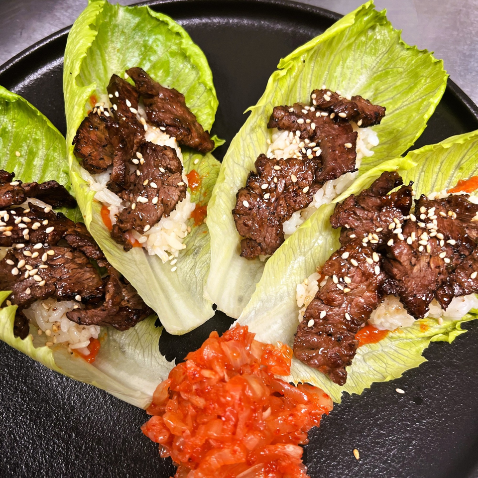 Four lettuce wraps filled with grilled meat, rice, sesame seeds, and a side of kimchi.