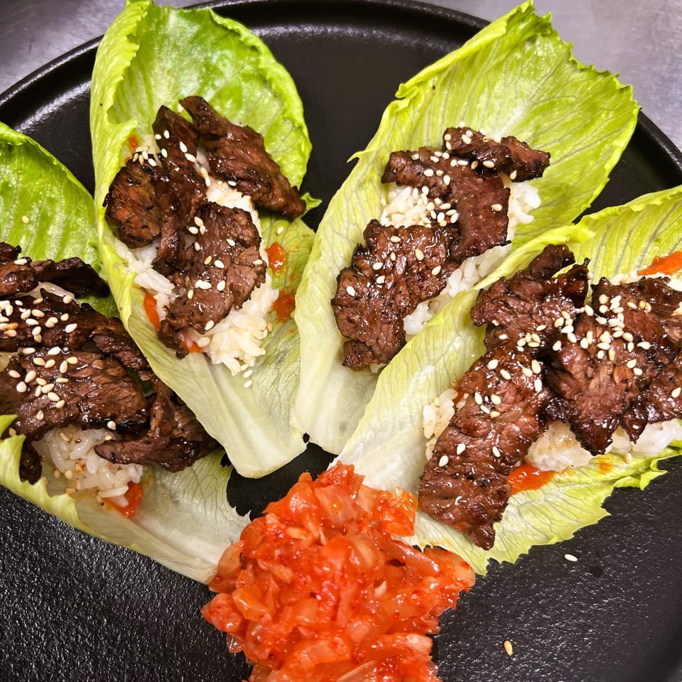 Four lettuce wraps filled with grilled meat, rice, sesame seeds, and a side of kimchi.