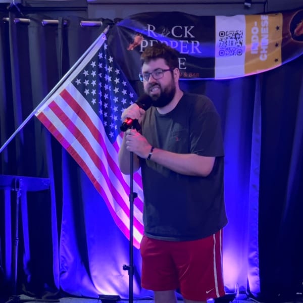 A man sings karaoke on stage with an American flag and Rock Pepper Fire banner behind him.