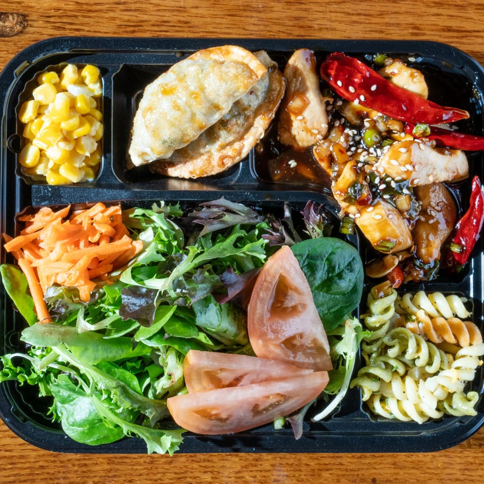 A Korean bento box with dumplings, spicy chicken, corn, salad, and pasta on a wooden table.