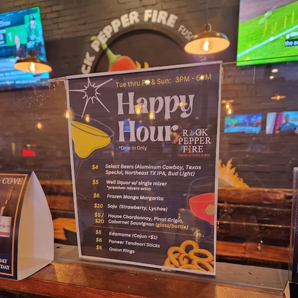 A Rock Pepper Fire Happy Hour menu board displays drink and appetizer specials in a restaurant setting.