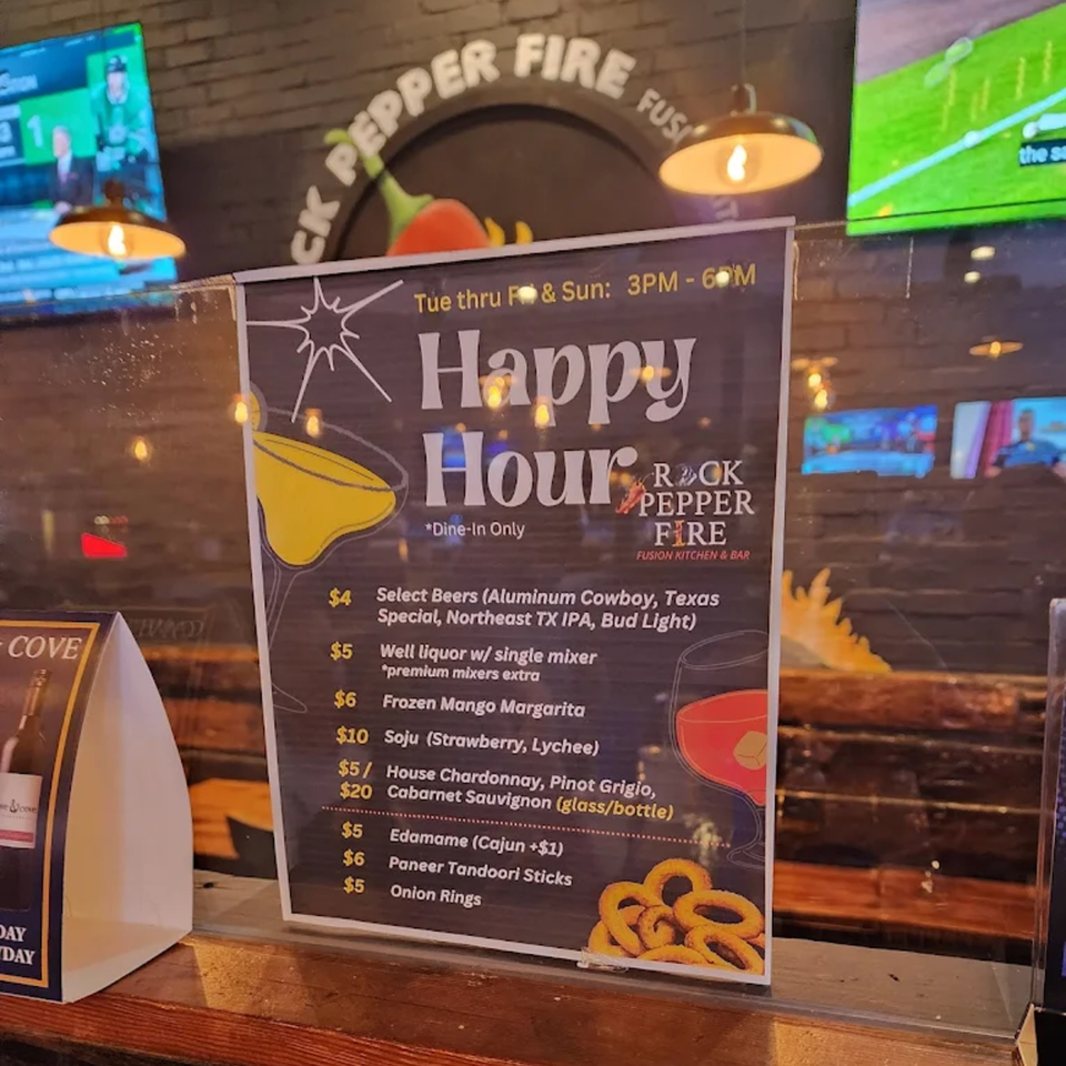 A Rock Pepper Fire Happy Hour menu board displays drink and appetizer specials in a restaurant setting.