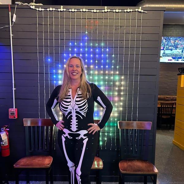 A woman in a skeleton costume smiles in front of a dark wooden wall with colorful string lights.