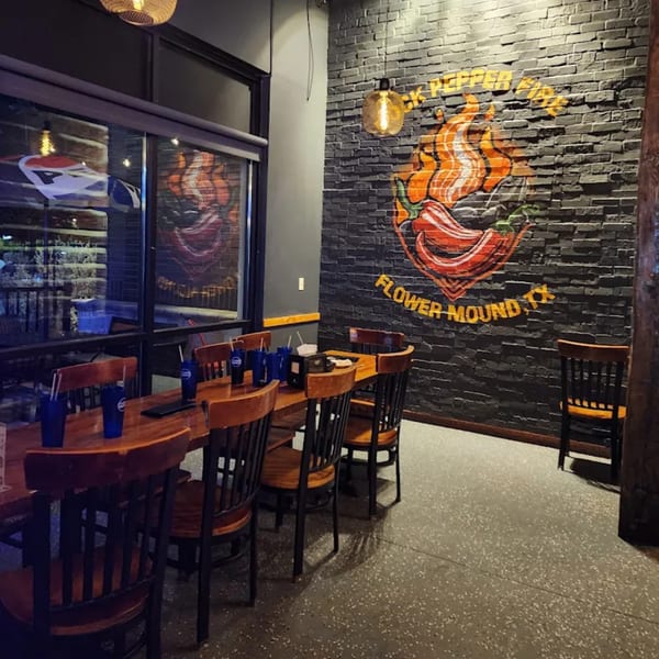 Interior of Rock Pepper Fire restaurant with a large chili pepper logo on a brick wall and wooden tables.