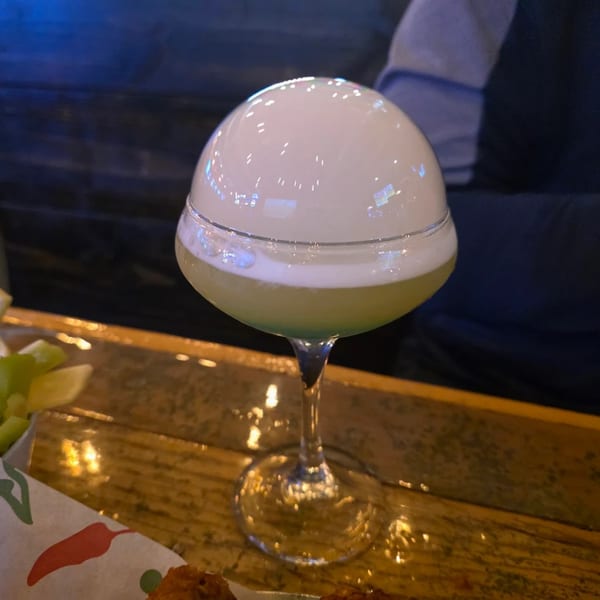 A green cocktail with a large, white foam dome sits on a wooden table at Rock Pepper Fire.